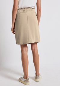 Jupe beige mi-longue avec une taille élastique, texture lisse et minimalisme dans les coutures, assortie à des baskets blanches et métalliques.