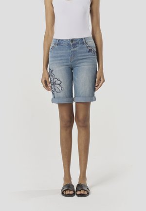 Jasnoniebieskie szorty jeansowe z podwiniętymi nogawkami, ozdobione kwiatowym haftem po jednej stronie. W komplecie dopasowany biały top na ramiączkach i czarne sandały.
