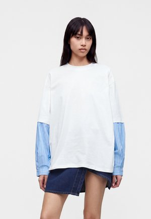 Jeune femme aux longs cheveux foncés portant une chemise blanche oversize superposée sur une chemise à manches longues bleue rayée et une jupe en jean foncé.