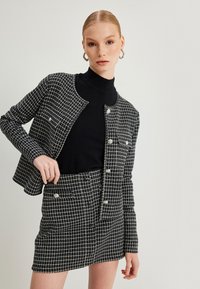 Sæt bestående af jakke og nederdel i sort-hvidt ternet mønster, med en struktureret design, tekstureret stof og sølvknap detaljer. Sort turtleneck nedenunder.