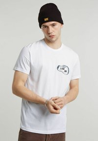 Giovane uomo che indossa una maglietta bianca G-Star Raw, un berretto di maglia nero e un braccialetto d'argento, con le mani giunte davanti a uno sfondo semplice.