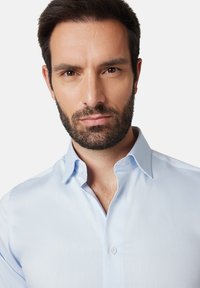 Jasnoniebieska koszula wizytowa z wyraźnym kołnierzykiem, gładką tkaniną, subtelną fakturą i guzikami. Zbliżenie koszuli noszonej przez modela.