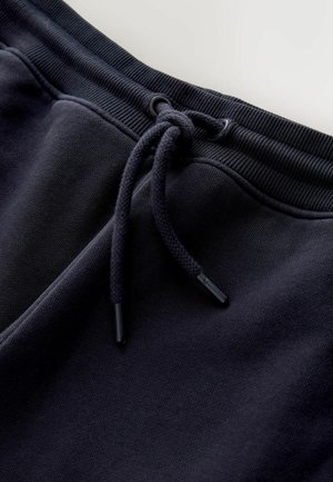Granatowe dresowe spodnie z elastycznego materiału mają szeroki pas, regulowany sznurek ze czarnymi końcówkami oraz gładką teksturę tkaniny.