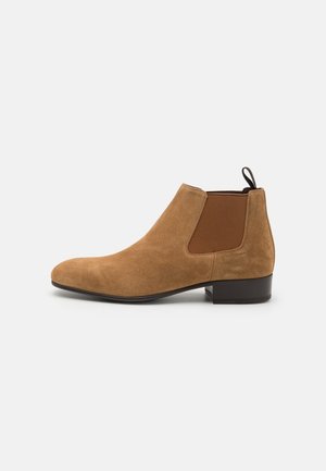Botine Chelsea din suede maro, cu panouri elastice laterale, vârf rotunjit și toc jos. Include o buclă pentru a facilita încălțarea.