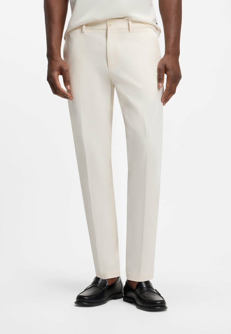 Homme portant un pantalon de costume crème slim fit et des mocassins en cuir noirs, debout sur un fond blanc uni.