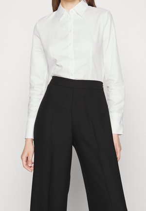 Kobieta ubrana w białą koszulę z kołnierzykiem zapinaną na guziki, wsuniętą w czarne spodnie z wysokim stanem i szerokimi nogawkami, stojąca na jednolitym tle.