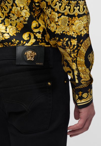 Pantalon noir avec une ceinture en cuir noire Versace ornée d’un emblème Méduse doré, associé à une manche et un poignet de chemise à motif floral noir et or.
