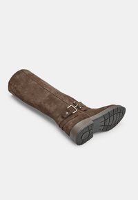 Stivale in suede marrone alto fino al ginocchio con suola piatta, caratterizzato da una fascia e un dettaglio in metallo con fibbia. La suola ha scanalature per migliorare la trazione.