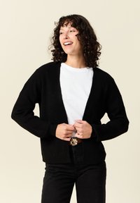 Svart stickad cardigan med öppen fram, avslappnad passform och långa ärmar, bärs över en vit t-shirt och kombineras med svarta byxor och ett bälte.