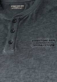 Dunkelgraues Baumwoll-Polohemd mit drei schwarzen Knöpfen, verziert mit einem gestickten Logo, das "STREET ONE MEN EXPERIENCE Unmatched Comfort" liest.