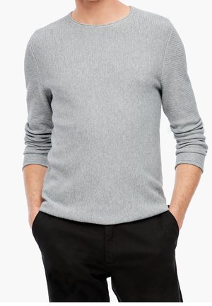 Homme les mains dans les poches, portant un pull léger gris texturé à manches longues et un pantalon noir sur fond blanc.