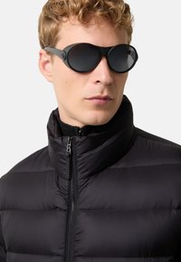 Mand med kort, krøllet blond hår iført sorte, runde solbriller og en sort pufferjakke med høj krave, der er lynet op.