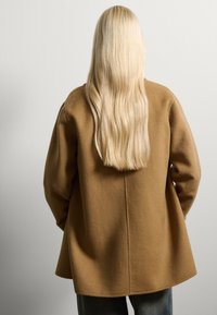 Cappotto di lana marrone con un fit rilassato, spalle cadenti e orlo posteriore più lungo. Tessuto liscio e dettagli di cucitura minimi.