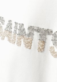 Tessuto bianco con un design di testo composto da perline argentate e simili a perle. Il testo presenta un effetto sfumato che va dall'argento a un beige chiaro.