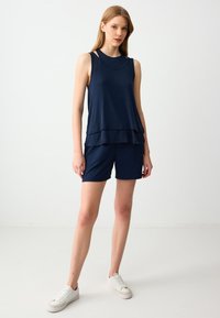 Vrouw in een gelaagde marineblauwe mouwloze top en marineblauwe shorts, staand met witte sneakers tegen een effen lichte achtergrond.