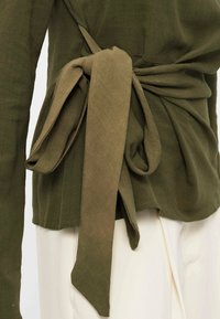 Close-up van een olijfgroene wikkeltop geknoopt in een zijstrik over een witte broek, met nadruk op de stoftextuur en het strikdetail.