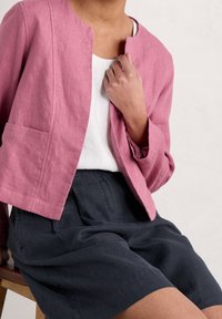 Ljusrosa linnejacka med rund halsringning, öppen framtill och långa ärmar. Bärs över en vit topp och mörkgrå shorts med veck.