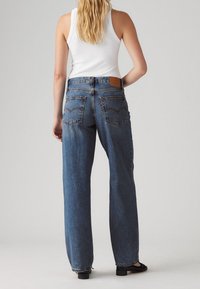 Femme aux longs cheveux blonds, portant un haut blanc sans manches, un jean bleu droit et des chaussures noires, debout, de dos.