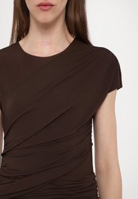 Brun, tettsittende topp laget av glatt, elastisk materiale; har drapert design med diagonale folder og korte ermer, som fremhever en slank silhuett.