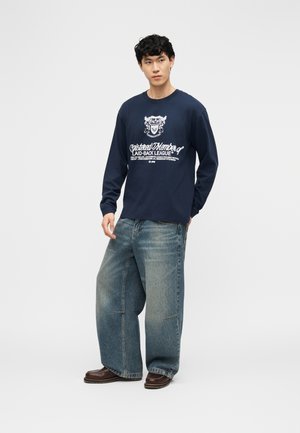 Νεαρός άνδρας που φοράει μπλε μακρυμάνικο μπλουζάκι με σχέδιο, φαρδιά ανοιχτόχρωμα τζιν παντελόνια και καφέ παπούτσια, στέκεται με το ένα χέρι στην τσέπη, μπροστά σε λευκό φόντο.