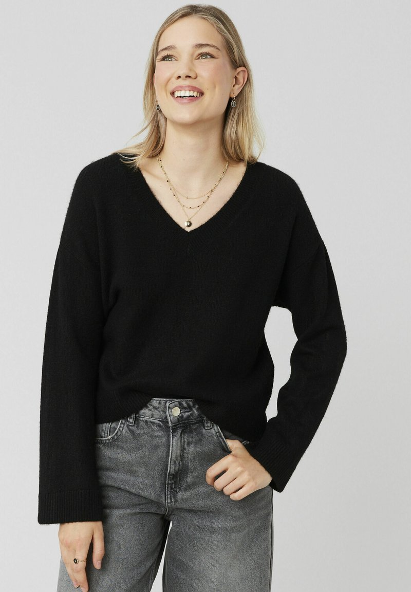 Pull noir à col en V avec des manches longues, fabriqué à partir d'un matériau doux. Associé à un jean gris taille haute, présentant une texture légèrement délavée.