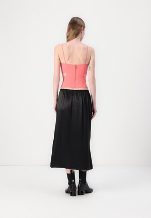 Vrouw met lang haar, van achteren gezien, draagt een koraalkleurige mouwloze top met uitsparingen, een zwarte midirok en zwarte laarzen met hak, tegen een witte achtergrond.