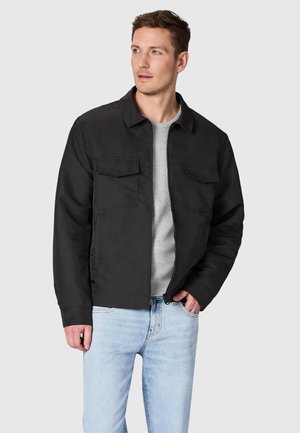 Man die een zwarte jas draagt over een grijze trui en lichtblauwe spijkerbroek, staat met één hand in de zak en kijkt naar de zijkant.