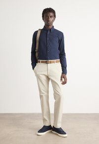 Marineknopen overhemden, beige broek, tan riem en marineblauwe sneakers. Glad stof, getailleerde pasvorm en horizontale stiksels op de schoenen.