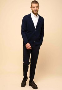 Marinblå stickad cardigan med sjalkrage, knappstängning, i kombination med smala svarta byxor och svarta snörskor, mot en beige bakgrund.