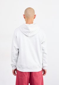 Glatzköpfige Person in einem weißen Hoodie und roten Hosen steht mit dem Rücken zu einer schlichten weißen Wand.