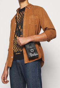 Bruine overhemd met knopen over een zwart graphic t-shirt, blauwe jeans, houdt een kleine zwarte leren crossbodytas vast met een band en sluiting detail.