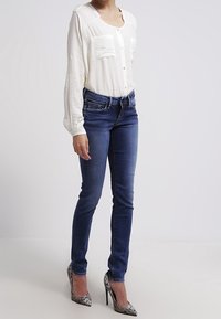 Blusa blanca de manga larga con tapeta de botones y dos bolsillos en el pecho texturizados, combinada con jeans ajustados de color azul oscuro y zapatos de tacón alto plateados.