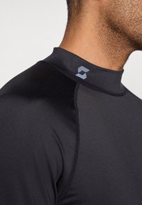 Μαύρο rash guard με ψηλό γιακά, απαλό ύφασμα και αντίθετο λογότυπο στο ύψος του λαιμού. Ολοκληρωμένος σχεδιασμός χωρίς ραφές βελτιώνει την άνεση και τη εφαρμογή.