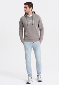 Jasnoszara bluza z kapturem z kieszenią z przodu i sznurkiem; zawiera duży nadruk tekstowy. Połączona z jasnoniebieskimi dżinsami dresowymi w stylu distressed i sportowymi butami.