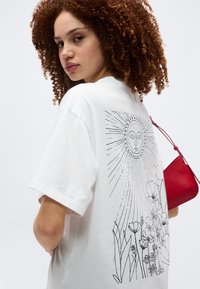 Γυναίκα με μπούκλες κόκκινων μαλλιών που φοράει λευκή μπλούζα με μαύρο σχέδιο σε γραμμή ενός ήλιου και λουλουδιών, κρατώντας μια κόκκινη τσάντα στον ώμο της.