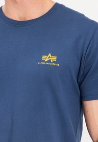 Blaues Baumwoll-T-Shirt mit einem runden Halsausschnitt, das ein gelbes Logo mit Streifen auf der linken Brust zeigt. Weicher Stoff, kurze Ärmel.