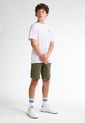 Weißes Baumwoll-T-Shirt, grüne Shorts, weiße Sneakers mit grünen Akzenten und blau-gestreifte Socken. Stehende Pose vor einem neutralen Hintergrund.