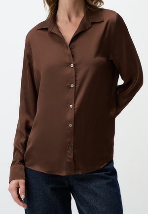 Femme portant une chemise boutonnée en satin marron à manches longues, associée à un jean bleu foncé, debout devant un fond blanc uni.