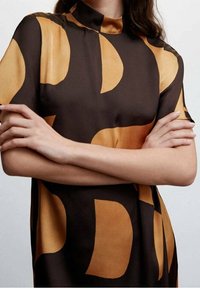 Blouse à manches courtes en tissu soyeux avec un motif géométrique audacieux brun et doré, dotée d'un col haut et d'une coupe décontractée.