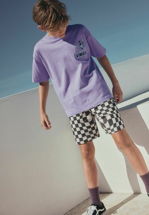 Boy wearing a lavender "Los Angeles Vibes" t-shirt, black-and-white checkered shorts, purple socks, and black sneakers standing outdoors.