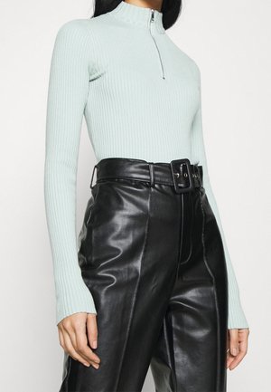 Femme portant un pull côtelé bleu clair avec une fermeture éclair sur le devant et un pantalon en cuir noir taille haute avec une large ceinture.