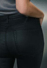 Crne traperice glatke teksture, s visokim strukom i dva stražnja džepa. Imaju detalje šavova i uski kroj.