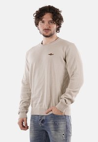 Maglione beige a girocollo in un morbido tessuto a maglia con polsini e orlo a costine; presenta un piccolo emblema ricamato di un'aquila sul petto.