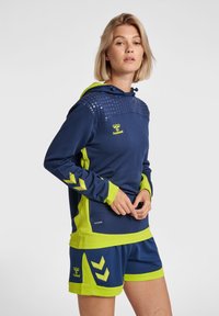 Felpa sportiva navy con accenti verde lime e punti testurizzati sulle spalle; abbinata a pantaloni corti coordinati con motivi a chevron.