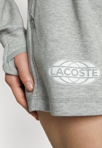 Sweatshirt cinza com textura suave, apresentando um grande logótipo branco "LACOSTE" na lateral, punhos canelados e detalhes de costura visíveis.