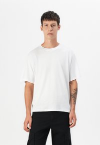 Hvid bomulds-T-shirt med korte, brede ærmer og rund halsudskæring. Har en afslappet pasform og sideslidser i kanten. Bæres sammen med sorte cargo-bukser.
