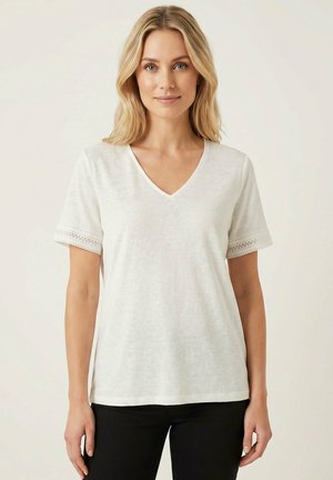 Mujer rubia con una camiseta blanca de manga corta y escote en V con borde de encaje en las mangas, de pie frente a un fondo claro y liso.