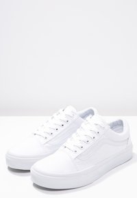Paire de baskets blanches basses avec lacets, détails de couture et semelles en caoutchouc sur une surface blanche avec un fond gris clair.