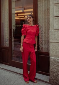 Completo rosso a due pezzi composto da un top con maniche a sbuffo e bottoni e pantaloni a vita alta, con gamba larga. Tessuto liscio, decorazioni minime.