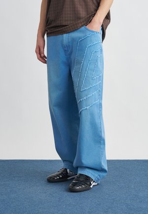 Oseba, ki nosi široke svetlo modre kavbojke z obrabljenim geometričnim vzorcem na stegnu, črne superge in rjavo karo srajco z roko v žepu.